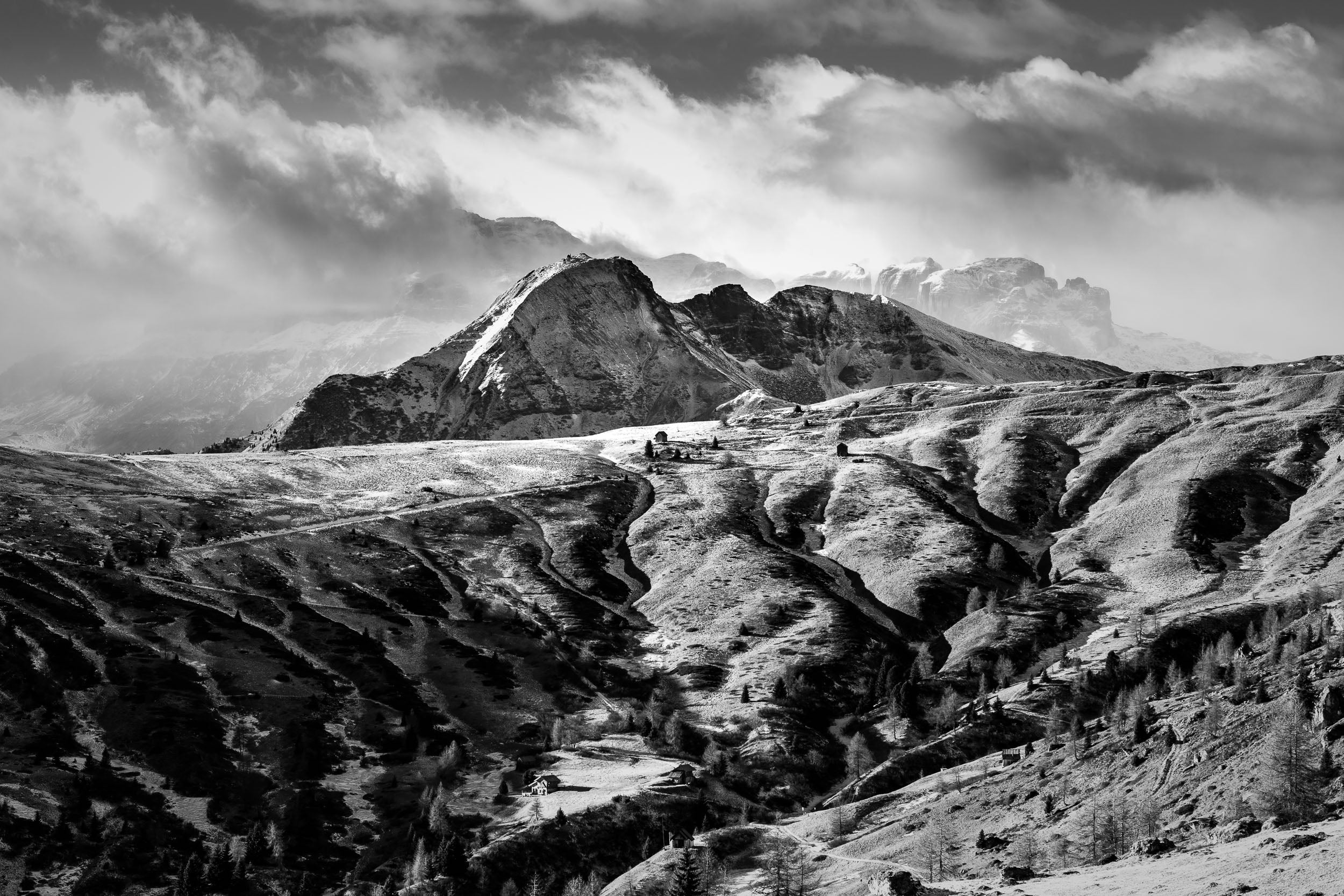 Fotografia in bianco e nero con vista autunnale sul Col di Lana. Sullo sfondo nuvole spazzate dal vento sopra il Gruppo del Sella. In primo piano alcune baite sui prati appena innevati.
