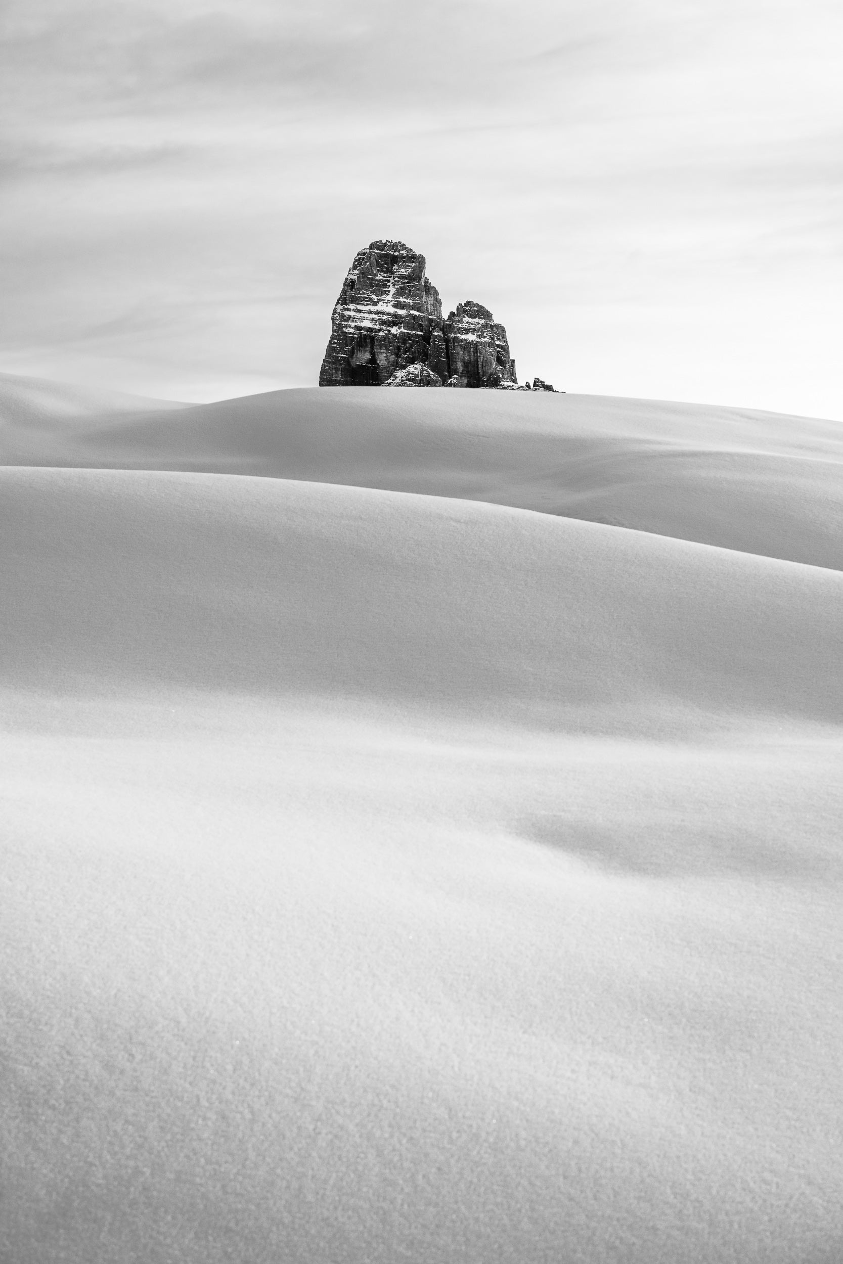Fotografia verticale invernale, le Tre Cime di Lavaredo spuntano in lontananza da una distesa di neve immacolata.