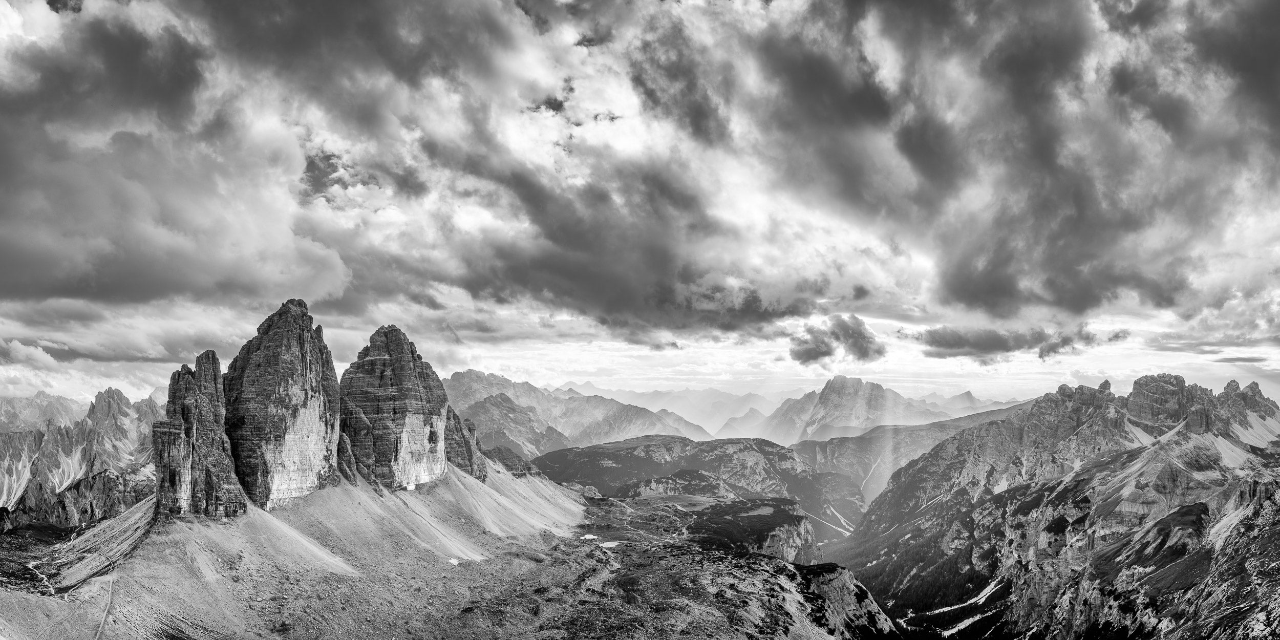 Fotografia panoramica in bianco e nero con vista drammatica su Tre Cime di Lavaredo, cielo molto nuvoloso con raggi di sole che illuminano alcune zone.